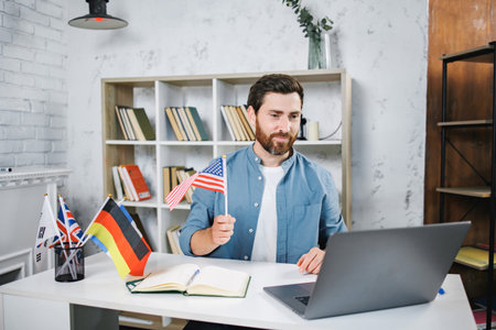 Bearded male tutor sitting at table with different flags and looking at laptopの写真素材