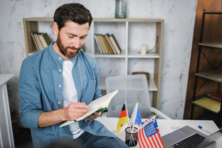 Bearded male tutor sitting on table with different flags and writingの写真素材