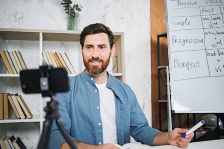 Bearded male tutor sitting at table next to whiteboard and recording a videoの写真素材