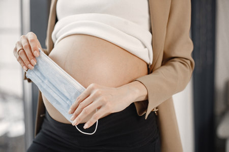 Midsection of pregnant lady with face mask standing near window in officeの写真素材