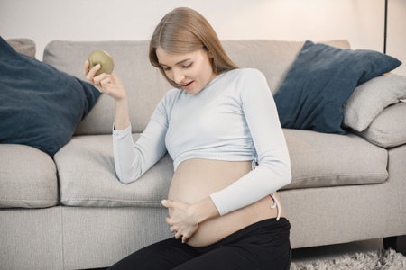Pregnant lady sitting near a sofa in living room holding an apple