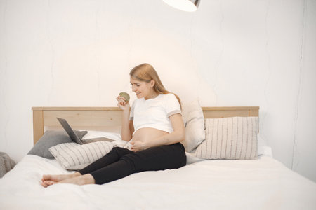 Pregnant lady lie on a bed in room and holding an apple while using a laptopの写真素材