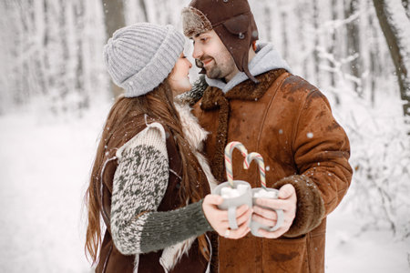 Romantic couple standing in winter forest, hugging and holding two cups with cocoa drink