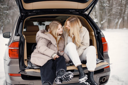 Blonde mother and teen daughter sitting in open cars trunk in winter forestの写真素材