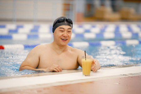 Asian man in hat and goggles drinking an orange juice in swimming poolの写真素材