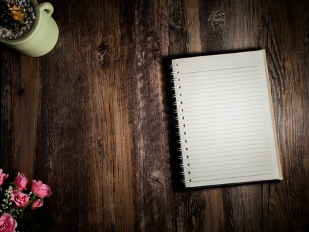 A beautiful notebook is placed on desk dark brown wood with a pink rose in the lower left corner. And a leaf in the top left corner.の写真素材