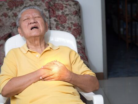 senile asian men sitting on a chair at living room with heart attacks.Both old man's hands on breast because of hard breathing, Face old man with pain caused by heart disease.の写真素材