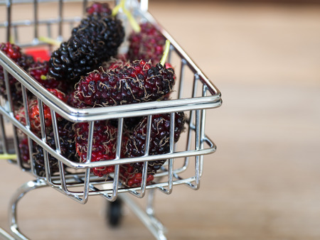 Close up group of mulberries in red shopping cart on wooden table. Mulberry this a fruit and can be eaten in have a red and purple color. Mulberry is delicious and sweet nature.の写真素材