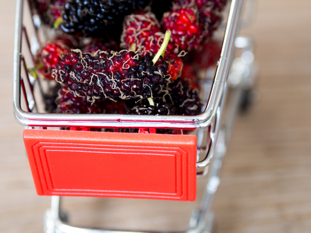 Close up group of mulberries in red shopping cart on wooden table. Mulberry this a fruit and can be eaten in have a red and purple color. Mulberry is delicious and sweet nature.の写真素材