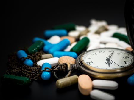 Antique clock on multicolored of drug and capsule is on the black background. Close up. We are against drugs (anti drugs), cure in container for health.Heap of green blue white round capsule pillsの写真素材