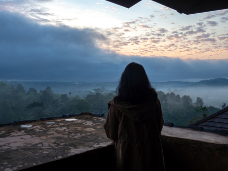 Women wear brown coat and standing on the balcony. And look outside with forest and mist in the morning. The golden sun is behind the clouds.の写真素材