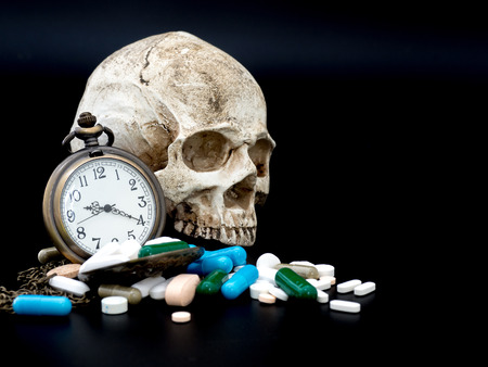 Antique clock on multicolored of drug and capsule behind clock is a skull. is on the black background. Close up. We are against drugs (anti drugs), cure in container for health.の写真素材