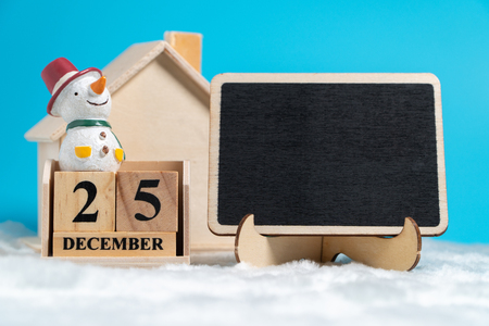 Snowman sitting on wooden block calendar set on 25 december beside blackboard on white wool in front of Wooden house. Copy space for text or content. Concept of Christmas day.の写真素材