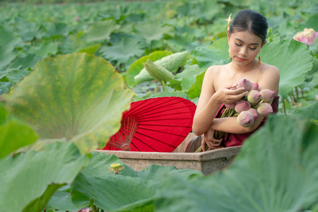 Beautiful asia women wearing traditional Thai dress and sitting on wooden boat with red paper umbrella in flower lotus lake. Her hands are holding a pink lotus. And her eyes are looking flowers.の写真素材