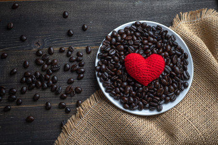 red heart on a lot of roasted coffee beans background on white plate on wooden table. Strong black...