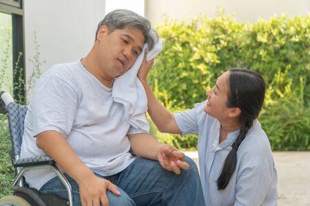 Middle-aged and obese men are paralyzed, Crooked mouth, Crinkled hands and Limb weakness  In the wheelchair and there is a wife using a towel on the face of her husband with love and good hopeの写真素材