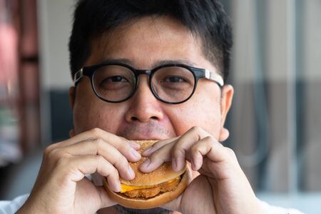 Asia man is eating hamburger in a fast food restaurant and enjoying delicious food. man in a White t-shirt and glasses holding a burger and enjoying the taste of hamburgerの写真素材