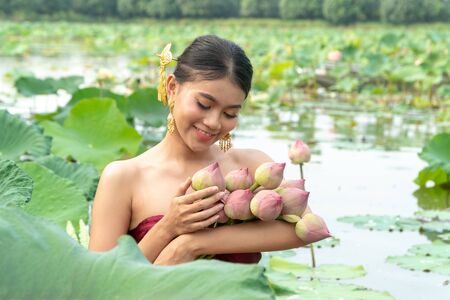 Beautiful asia women wearing traditional Thai dress and sitting on wooden boat in flower lotus lake. Her hands are holding a pink lotus and picking up flower.  And her face is be happy and smile.の写真素材