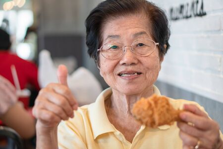 Asian elderly women are eating fried chicken. In the restaurant And lift the thumb. Her smile and look happy. Concept of aging society, Preparedness After retirement And living a happy life.の写真素材
