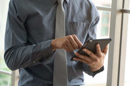 Happy young business man using digital tablet and smiling, standing near the window in office