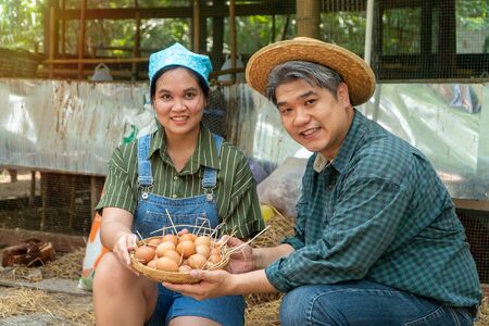 Asian couple farmer holding fresh chicken eggs into basket was sitting near hen beside chicken farm.Smiling because happy with the products from the farm. Concept of Non-toxic foodの写真素材