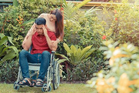 Asian Man in a wheelchair and Unhappy and painful. A woman standing behind the wheelchair and is encouraging her husband, whose feet hurt his leg due to an accident. Concept of caring and supportの写真素材