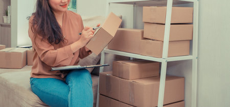 Young entrepreneur, sitting on the sofa and working at home. She smiles and holds a packing box for...