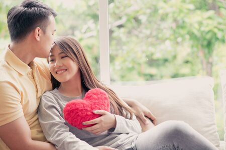 Asian Couples Sitting on the sofa In which women Holding a red heart And smiling happily. Concept of expressions of love and warmth for lovers, Valentine day.の写真素材