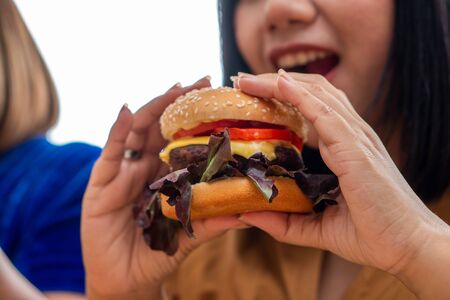 Hungry overweight woman smiling and holding hamburger and sitting in the living room, her very happy and enjoy to eat fast food. Concept of binge eating disorder (BED).の写真素材