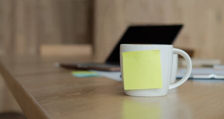 A white coffee cup with a blank yellow Sticky Note on the cup. Resting on the desk and laptop is placed on the back.の写真素材