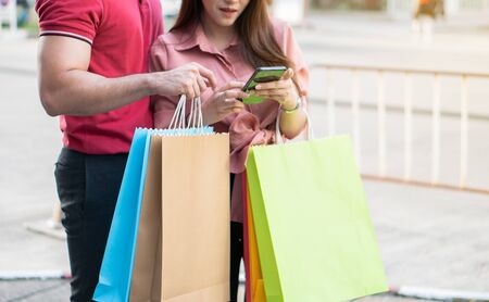 Happy young couple of shoppers walking in the shopping street towards and holding colorful shopping bags in hand and use a smartphone for check promotion. Concept of sale and Black Friday shoppingの写真素材