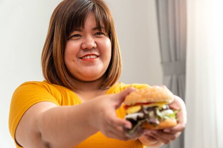 Hungry overweight woman smiling and holding hamburger and sitting in the bedroom, her very happy and enjoy to eat fast food. Concept of binge eating disorder (BED).の写真素材
