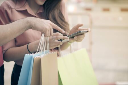 Happy young couple of shoppers walking in the shopping street towards and holding colorful shopping bags in hand and use a smartphone for check promotion. Concept of sale and Black Friday shoppingの写真素材