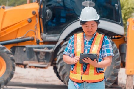 Woman Engineering wearing a white safety helmet standing In front of the backhoe And are using tablet to check the blueprint with constructionの写真素材