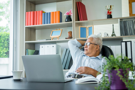 Asian elderly man sitting in front of a laptop computer, He is sitting back in his chair and relax....