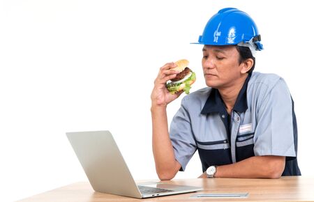 Asian man worker smiling and holding a hamburger in the living room While working from home, hem very happy and enjoy to eat fast food. Concept of binge eating disorder (BED).の写真素材