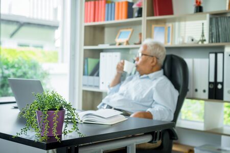 Asian elderly man sitting in front of a laptop computer, He is sitting back in his chair and relax. The elderly can still work And create sentences for society, virus quarantine,の写真素材