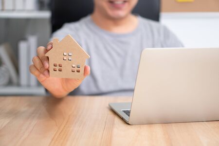 Smiling businessman holding a wooden house in home workspace. The concept of working from home until the end of Coronavirus (COVID-19) quarantine, happily freelance working lifestyleの写真素材