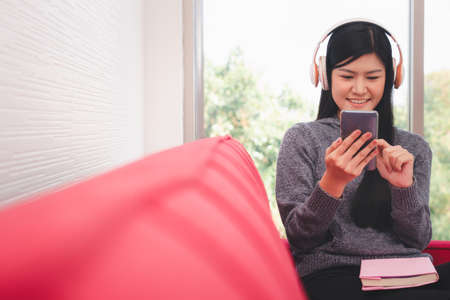 Cute Asian woman is sitting on the sofa in the morning And send messages on mobile phones While listening to music And with a book on the lap In the living roomの写真素材