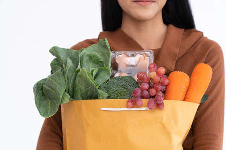 Happy Asian woman is smiling and carries a shopping paper bag after the courier from the grocery came to deliver his goods at home. Concept of Supermarket delivery for a new lifestyleの写真素材
