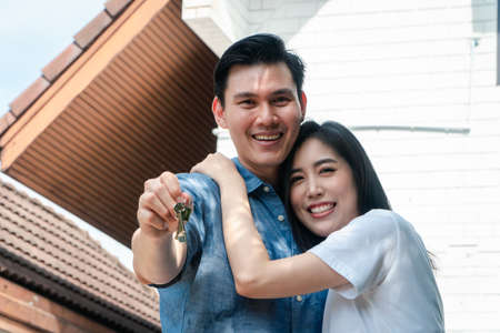 Happy smiling couple in front of the home and holding keys to New Home On Moving In Day, and start a new life family. Concept of homeowner and relocationの写真素材