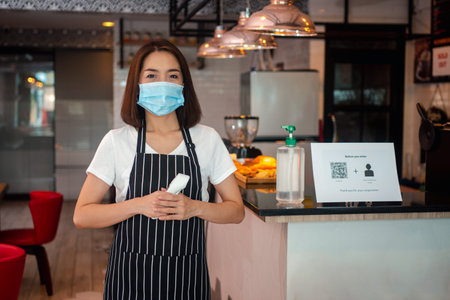 Asian waitress woman wearing face masks and holding an infrared forehead thermometer to check body...