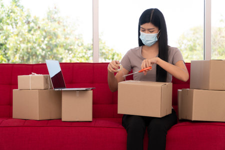 Happy young Asian entrepreneurs on a mask are packing boxes for delivering products to customers....