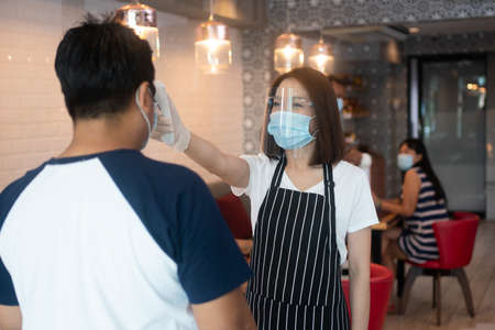 Asian waitress woman wearing face masks and holding an infrared forehead thermometer to check body temperature for virus symptoms of customers before entering the restaurant ( coffee shop ).の写真素材