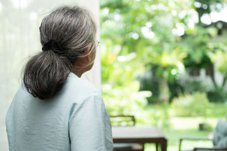 A sad old elderly woman uses Walker for standing in front of windows and looking outside and feeling lonely. Concept of depression caused by illness and Elderly caregiversの写真素材