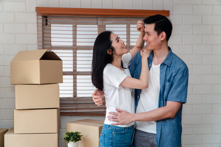 Smiling young Asian happy couple hugging boyfriend with cardboard boxes at moving day in their new...