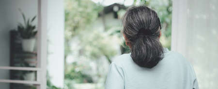 A sad old elderly woman uses Walker for standing in front of windows and looking outside and feeling lonely. Concept of depression caused by illness and Elderly caregiversの写真素材