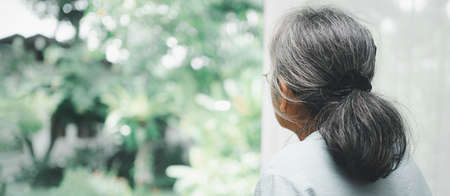 A sad old elderly woman uses Walker for standing in front of windows and looking outside and feeling lonely. Concept of depression caused by illness and Elderly caregiversの写真素材