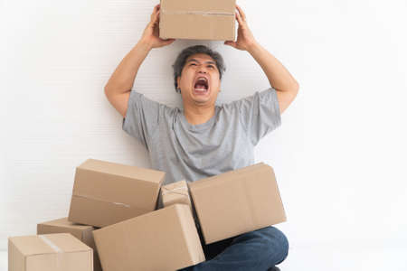 Asian Shopaholic man Shocked and sitting on the floor in the living room and Cardboard Box on top of him after The courier delivered to the home. Concept of shopping online and Shopaholic.の写真素材
