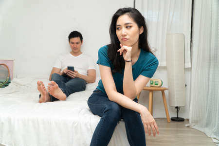 Unhappy Asian woman in bed and feeling Bored and grumpy her husband because tired of her husband who ignores her and uses the smartphone all time. Concept of relationship problem from technologyの写真素材